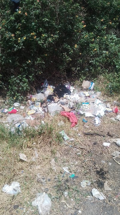 The scene depicts an outdoor area in Nkaimurunya ward, Kenya, characterized by a significant accumulation of mixed refuse at the base of dense green vegetation. A substantial pile of garbage, predominantly in the midground, includes numerous plastic bags, cardboard, scattered wrappers, two distinct red fabric items, and a dark, possibly charred, mass of material. The ground in the foreground consists of dry, patchy grass and exposed soil. The background features a thick bush or hedge with dark green leaves and small yellow and pinkish-orange flowers. The area is illuminated by natural sunlight, casting subtle shadows. No individuals are visible.