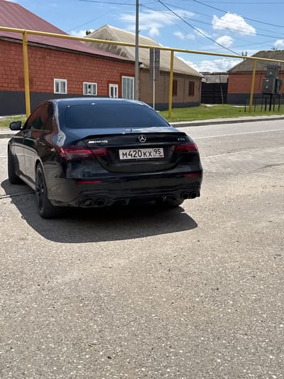 A black Mercedes-Benz E53 AMG is parked on a paved road in Mayrtup, Russia. The car has a license plate that reads "M420KX 95". In the background, there are several brick buildings with tiled roofs and a yellow pipe. The sky is blue with some clouds. The scene is a typical street view in a Russian town.