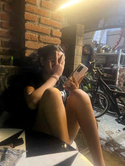 A young woman sits in what appears to be a dimly lit, outdoor cafe or seating area. She is looking at her phone, holding it in her right hand. Her left hand is covering her face. She is wearing a black t-shirt and has bracelets on her left wrist. Behind her, a brick wall is visible. In the background, there are bicycles, shelving units with various items, and an umbrella. The scene suggests a casual, possibly cluttered environment. The location is identified as Mendoyo, Indonesia.