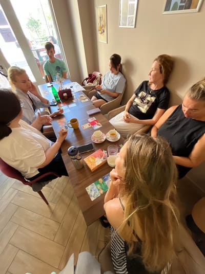 A group of seven individuals, appearing to be adults, are seated around a wooden table in what appears to be a cafe or indoor seating area, possibly located in Donji Pobori, Montenegro, given the context of the request. The group is engaged in playing a card game. Visible on the table are playing cards, cups of tea or coffee, a bottle of green liquid, and a thermos. The individuals are dressed casually. Sunlight streams in from a large window or glass door on the left side of the frame, illuminating the scene. The floor is tiled in a herringbone pattern.