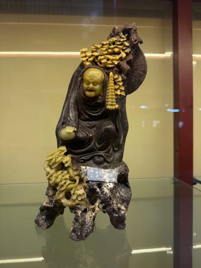 A carved stone sculpture of a monk is displayed in a glass case. The monk, with a yellow face and dark robes, is seated on a textured, dark brown base that resembles rock formations and tree roots. The sculpture incorporates lighter, yellowish-green carved elements that appear to be organic, such as clusters of small leaves or flowers, and a miniature pagoda. A small metal plaque with inscriptions is affixed to the base. The background of the display case is a pale yellow wall with horizontal fluorescent lighting above. The sculpture is positioned on a reflective green surface. There are no people or specific geographical landmarks of มาบฟักทอง, Thailand visible in the image.