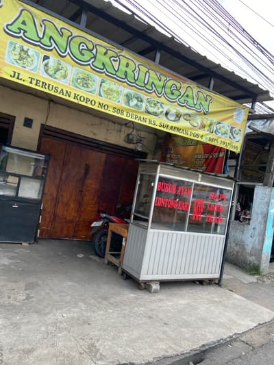 The image shows a street food stall in front of a storefront, located at JL. Terusan Kopo No. 508, Kecamatan Margahayu, Indonesia. The stall is a small metal cart with a glass front and red lettering advertising "Bubur Ayam," "Lontong Kari," and "Sapi Kambing." The stall is parked on a sidewalk, next to a motorcycle parked in front of the storefront. The storefront has a large yellow banner above the entrance, which reads "Angkringan" in green lettering. The banner also includes several pictures of food items and a phone number. The storefront has a wooden door and a window. The scene is captured from a low angle and shows a grey concrete sidewalk. The environment is bustling with activity, although no people are present.  The building is a single story and appears to be a shop or a restaurant.