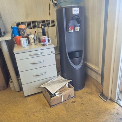 In Nowa Wieś Książęca, Poland, this image captures a cluttered indoor scene, likely a utility room or office space. A large, dark grey Culligan water cooler stands prominently on the right side of the frame. To its left, a white filing cabinet with silver handles is positioned, holding several items on its top surface. These include a blue bag of "CUKIER" (sugar), an orange mug, a white mug with "+ 100" written on it, a black and white mug adorned with paw prints, and a mug featuring an image of a town and the word "EMILIA". Behind the filing cabinet, a white electric heater with horizontal slats is visible. A glass electric kettle containing water sits on the filing cabinet's surface. In the foreground, a cardboard box is open, containing various items, including a crumpled sheet of paper with a printed grid and a metal handle. Rolls of tape are stacked near the filing cabinet. The floor is covered in a speckled yellow linoleum. The walls are paneled with vertical white strips, and a door frame is visible on the far right. The overall impression is one of functional disarray.