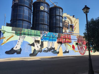 The image shows a mural painted on the side of a building in Valencia de Alcántara, Spain. The mural features a series of large, dark silos behind a colorful painting of legs and feet wearing traditional Spanish attire, including patterned stockings and black shoes. Above the legs, a portrait of a man with a beard is painted, along with wheat stalks and poppies. To the right of the building, a lamppost stands next to a leafy tree. The scene is illuminated by natural daylight, casting shadows on the asphalt road in the foreground.
