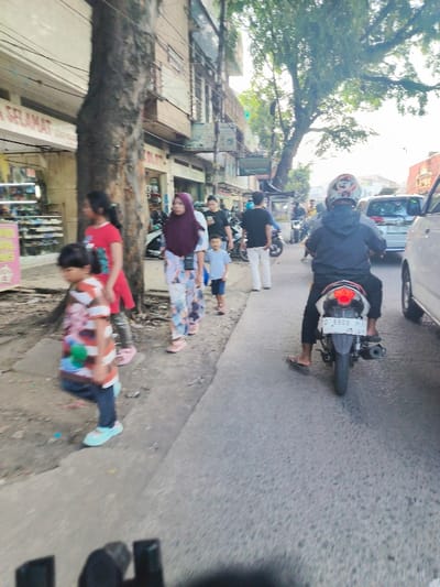 The image shows a street scene in Kecamatan Dayeuhkolot, Indonesia. Five people are walking on the sidewalk on the left side of the image. A man in a blue shirt is leading a small child. A woman wearing a red shirt and a young girl are walking next to each other. A woman wearing a hijab is walking behind them. On the road, a man on a motorcycle is riding towards the viewer. A white van is parked on the right side of the image. The road is paved and there are buildings on both sides of the road. The buildings are painted in various colors, such as beige, light blue, and red. There are trees on the left side of the image. The sky is overcast and the image is taken on a cloudy day. The image is taken from the perspective of someone riding on a motorcycle. The license plate on the motorcycle reads "D 6908 NIA".