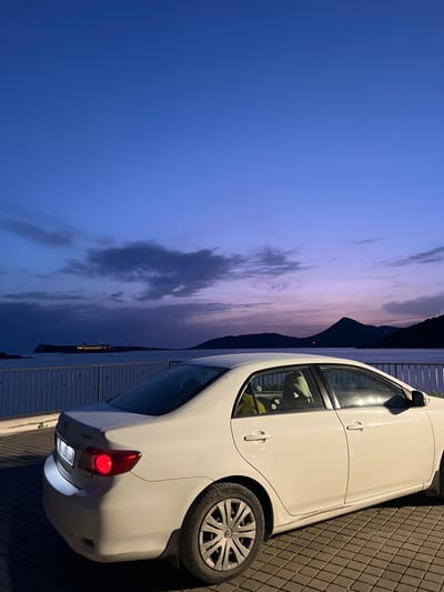The image presents a white car parked on a paved surface, likely a sidewalk or parking area in Luštica, Montenegro. In the background, a body of water stretches to the horizon, reflecting the colors of the twilight sky. The sky is characterized by gradients of blue and purple hues, with scattered clouds. In the distance, a dark silhouette of a mountain or hill is visible against the horizon. The lighting suggests it is either dusk or dawn. The car's rear lights are illuminated, and the details of the wheels and body are apparent. A metal fence lines the edge of the paved area, separating it from the water.