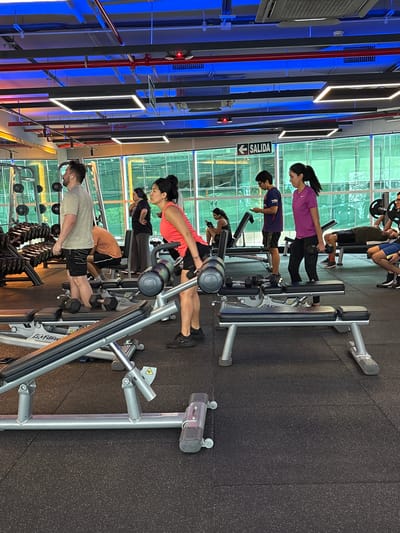 The image depicts the interior of a modern gym located in San Isidro, Peru, with large windows revealing a cityscape in the background. The gym features a dark rubber floor and a high ceiling with industrial lighting, including blue neon accents. Several individuals are engaged in various exercises. In the foreground, a woman in a pink tank top and black shorts is performing dumbbell rows. Adjacent to her, a man in a gray t-shirt and black shorts stands near a rack of dumbbells. Further back, other gym-goers are visible: a person in a purple t-shirt is holding a dumbbell, another individual in a blue and purple shirt is looking at their phone, and a person is lying on a bench performing a bench press with weights. The gym is equipped with weight racks, benches, and other exercise machines. The overall atmosphere is one of activity and fitness.