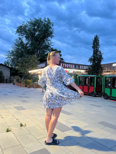 This media file depicts a woman standing on a paved walkway in what appears to be an outdoor entertainment area. She is wearing a light-colored dress with a brown, leaf-like pattern and dark sandals. Her hair is blonde, and she is wearing sunglasses on her head. The woman is holding the hem of her dress up with her left hand, and her pose suggests she is looking to her left.
In the background, there is a colorful, toy-like train, primarily red and green, with illuminated outlines. Adjacent to the train is a building with signage. Visible text on the building reads "КУРОЧКИ & БУЛОЧК" in large, illuminated letters. This suggests a food establishment.
The scene is set outdoors, with trees and a cloudy sky visible. The lighting suggests it is late afternoon or early evening, with shadows cast on the ground. The sky is a deep blue with some white clouds, indicating possibly overcast or twilight conditions. The paved walkway is made of rectangular stones in varying shades of gray and beige, with some small weeds growing in the cracks. The overall atmosphere appears to be relaxed and casual, possibly a promenade or a fairground. The location is identified as Shchelkino urban settlement, Ukraine.