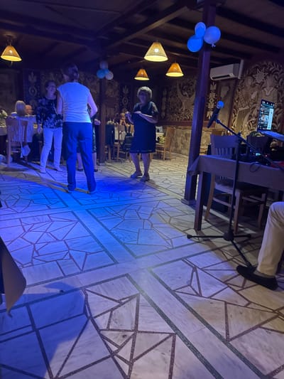 The image depicts an indoor gathering, possibly a party or restaurant, with a dance floor. Several individuals are present, some of whom appear to be dancing. The lighting is predominantly blue, cast from above, illuminating the patterned tiled floor. Decorative elements include lamps hanging from the wooden ceiling and a mural on the stone wall. Balloons are visible near the ceiling, suggesting a celebration. A table with a microphone and a laptop is set up, indicating potential entertainment or a DJ. The overall atmosphere appears lively. The location is identified as Sevlievo, Bulgaria, suggesting this is a scene within the city.