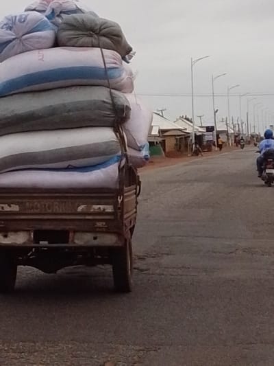 An open-back truck overloaded with sacks travels on a paved road in Dungu, Ghana. The truck's tailgate reads "MOTOR KING". A person on a motorbike rides in the same direction, further down the road. Along the road, houses and streetlights are visible. Pedestrians are also present.