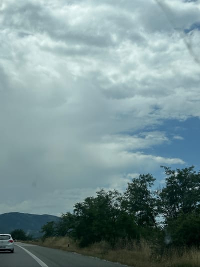 An outdoor daytime scene depicts a partially cloudy sky transitioning from overcast grey on the left to patches of blue with white clouds on the right. The image captures a view from a moving vehicle on an asphalt road in or near Tserovo, Bulgaria. A white hatchback car, bearing the license plate "CB 46 14 HT", proceeds ahead on the left lane, its rear visible. The road features a solid white shoulder line and a dashed white lane divider. Along the right side of the road, dry, golden-brown grasses and shrubs are present, punctuated by small red and white roadside markers. Further right and extending into the midground, a dense line of deciduous trees with dark green foliage borders the road. In the background on the left, a range of large, verdant mountains or hills is visible beneath the heavier cloud cover.