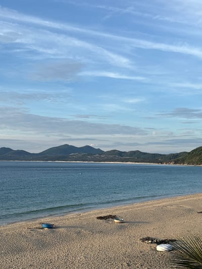 The image depicts a serene coastal landscape in Song Cau, Vietnam. A calm, blue ocean meets a sandy beach lined with sparse vegetation and a few small boats. In the background, a range of green, forested mountains stretches across the horizon under a partly cloudy sky. The scene is quiet, with no people visible. The lighting suggests it is either early morning or late afternoon, casting soft shadows on the sand.