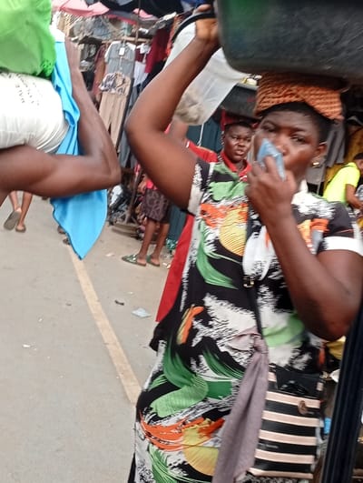 A woman in a floral print dress balances a large container on her head in a bustling market. She has a striped purse slung across her shoulder and is holding something to her nose. Another person carries bundles of fabric nearby. Clothing hangs on racks behind her, and other shoppers are visible in the background, adding to the busy atmosphere of the market.