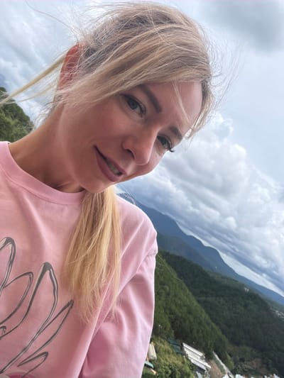 The image is a portrait of a woman with blonde hair, wearing a pink sweatshirt with a large, abstract design on it. She has a slight smile, and her hair is being blown by the wind. In the background, there is a cloudy sky and a view of a lush green mountainside with some buildings visible on it. The location is identified as Xuân Thành, Vietnam.