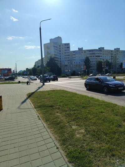 This media file captures a sunny daytime scene on a street in Salihorsk, Belarus. The foreground features a paved sidewalk with a grassy verge, casting a long shadow from a nearby lamppost. A person is seen walking with a suitcase on the sidewalk, suggesting travel or movement.
The street itself is busy with traffic, including several cars and what appears to be a dark-colored station wagon in the right lane. Traffic lights are visible, indicating an intersection or junction. The background is dominated by a series of large, multi-story apartment buildings, typical of Soviet-era architecture. These buildings are light-colored, with many windows and balconies.
The sky is a clear blue with a few scattered white clouds, suggesting fair weather. The sunlight indicates it is likely midday or early afternoon. A billboard is visible on the left side of the street, but the text is too small to read clearly. The overall atmosphere is one of everyday urban life on a bright, clear day.