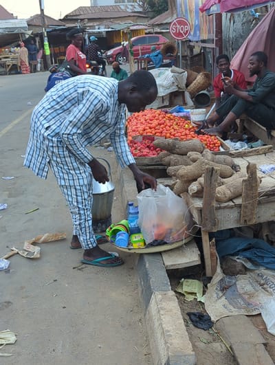 The image depicts a street scene in Shaba, Nigeria. A man in a blue and white striped shirt and pants sits on the ground next to a tray. The tray contains water bottles, small plastic cups, and a clear plastic bag with unknown contents. Next to the tray is an assortment of yams and tomatoes. Behind the tomato stand, two men are sitting and looking at a cell phone. A “STOP” sign is mounted nearby. In the background, other people are visible along the street with motorcycles and cars parked. The environment appears to be an open-air market area on a dirt and concrete surface.