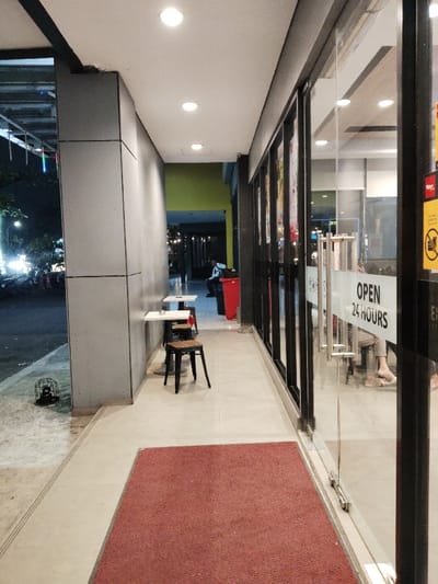 The scene is a narrow corridor with tiled floors and a red carpet. It is located in the city of Bekasi Regency, Indonesia. There are glass doors on the right side leading into a shop with "OPEN 24 HOURS" sign. Two white tables with two chairs are placed on the left side of the corridor. One person is sitting at a table in the distance. There are lights on the ceiling of the corridor. Outside the corridor, through the glass doors, it is nighttime, and there are streetlights.