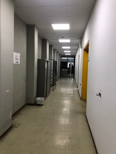 The image shows a long, narrow hallway with grey walls and a grey tiled floor. The hallway is lit by overhead fluorescent lights. There is a grey metal cabinet on the left side of the hallway, and a yellow door on the right. A trash can is in the middle of the hallway. At the end of the hallway, there is a door with a window. The image was taken in Agia Paraskevi, Greece, at the coordinates 37.998, 23.82. No people are present.