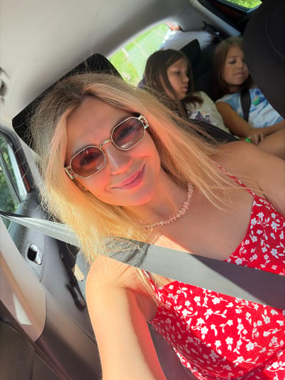 In a vehicle, possibly on a road trip, a woman with blonde hair and sunglasses smiles at the camera. She is wearing a red floral dress and a beaded necklace. In the background, two young girls are seated in the back of the car. The woman's face is partially illuminated by sunlight. The car's interior, including a seatbelt, is visible. The setting does not provide specific details to confirm a location in Bare Kraljske, Montenegro.