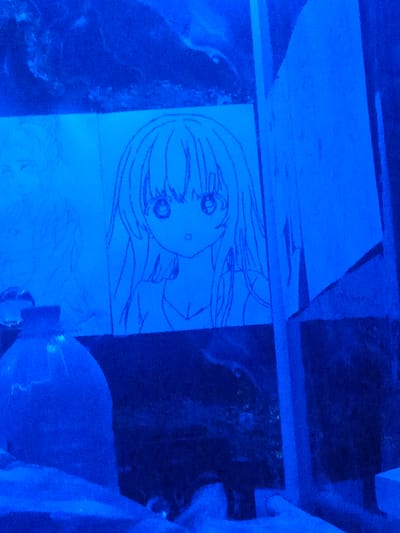 In the city of Yangi Chinaz, Uzbekistan, an image depicts a display of hand-drawn sketches on paper, illuminated by blue light. The primary focus is a sketch of an anime-style girl with large eyes and long hair, rendered in dark lines. To the left, another partial sketch of a person's head and upper body is visible. In the foreground, a large, translucent blue bottle with a black cap stands, appearing to be made of plastic. The background consists of a dark, textured surface, possibly a wall or display material, also bathed in the pervasive blue light. The overall scene suggests an artistic display or an exhibition, with the sketches prominently featured.