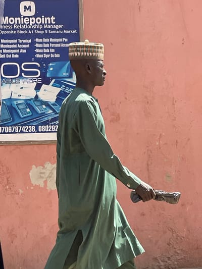 A male subject is captured in profile, walking from left to right, in an outdoor setting in Samaru, Nigeria. He wears a light olive-green long-sleeved tunic and matching trousers, topped with a round, patterned hat featuring a central knob. His right hand holds a dark, irregularly shaped object, possibly wrapped in black material. The background consists of a reddish-orange building wall exhibiting significant paint wear and discoloration. To the left, a large blue and white commercial sign for "Moniepoint Business Relationship Manager" is partially visible. The sign specifies the location as "Opposite Block A1 Shop 5 Samaru Market" and advertises services including "Moniepoint Terminal," "Moniepoint Account," and "Moniepoint ATM," displaying graphics of POS devices and a Verve card logo. The scene is brightly lit by natural sunlight.