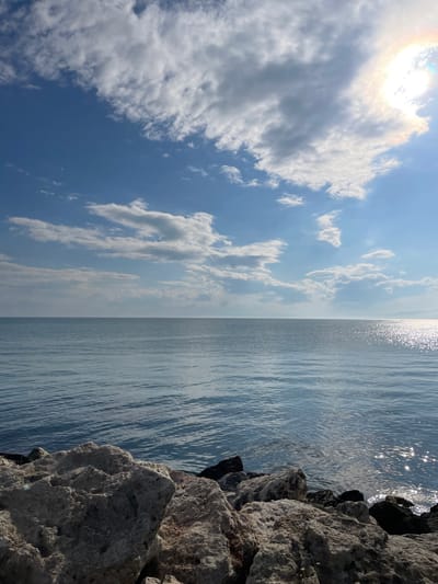 The image shows a seaside scene, divided roughly in half vertically. The left half of the image features a blue sky with numerous white and gray clouds, with the sun visible in the upper left corner. The right half shows water with sunlight reflecting off the surface. A rocky shoreline is visible along the right edge. The location is likely Kavarna, Bulgaria, given the rocky coastline and the appearance of the Black Sea.