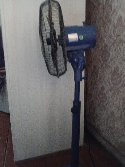 The image shows a blue oscillating fan standing on a dark floor next to a textured, patterned wall. The fan is positioned in front of a plain, light-colored surface, possibly a wall or screen, which occupies the left side of the frame. The fan has a protective grill covering its blades and a central motor housing with a label and a green circular sticker. The stem of the fan is also blue, and it sits on a circular base. The lighting in the scene appears to be dim, casting shadows. There are no people visible in the image. This scene does not provide any information to determine its location within the city of Kamen-na-Obi, Russia.