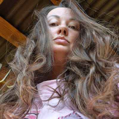 A close-up, low-angle shot shows a young woman's face from a perspective looking upwards, with her long, wavy, brown and blonde hair spread out around her. She is wearing a pink shirt with decorative, striped trim. The background consists of a corrugated, dark brown surface and a wooden plank. The lighting is soft and diffused, suggesting it might be daytime, possibly indoors or outdoors in diffused light. The woman's expression is neutral. The image has a slightly tilted orientation.
