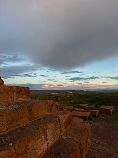 The image depicts a landscape view from the ancient city of Ani, located in present-day Armenia, with focus on the weathered stone ruins of the site and a dramatic sky above. In the foreground, a fragment of a structure, possibly a wall or a part of a building is constructed from reddish-brown stones with visible lichen and moss. The blocks are stacked unevenly. Beyond the wall, the landscape slopes downward into a valley with green grass. In the midground, there are a few buildings. The sky takes up a significant portion of the image, transitioning from dark, stormy clouds on the left to a softer, pastel-colored sunset gradient. The atmosphere suggests either early morning or late evening light conditions. There are no people or animals visible in the frame.