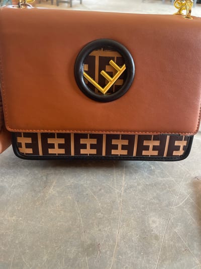 This media file showcases a close-up view of a brown handbag with a distinctive "FF" logo in gold, set against a patterned strip of the same logo in brown and beige. The bag appears to be placed on a textured, grey surface with visible scratches and markings, suggesting an indoor setting, possibly a workshop or a storage area. The lighting is neutral, indicating it could be daytime or artificial lighting, with no strong shadows. The image focuses on the details of the handbag, highlighting its material texture and the logo's metallic sheen. There is no visible text in the image. The context suggests this item might be related to fashion or retail, possibly being presented or stored in Nigeria.