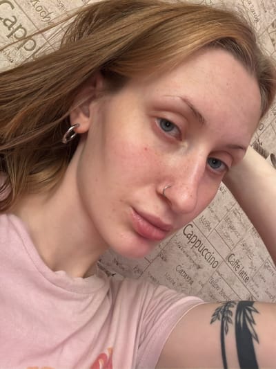 The image shows a close-up, selfie-style portrait of a young Caucasian woman with long, reddish-brown hair, fair skin with freckles and a slight redness on her cheeks. She has blue eyes and is wearing a small silver hoop earring in her left ear and a small silver hoop piercing in her nose. She is wearing a light pink t-shirt. The background appears to be a wall with a repeating pattern of coffee-related words like "Cappuccino," "Mocha," "Caffè latte," and various coffee bean origins, suggesting a cafe or a themed environment. Her right arm is propped up behind her head, resting on the patterned surface. There is a black tattoo of palm fronds visible on her forearm. The lighting is soft and seems to come from above and slightly to the left, casting gentle shadows. The image quality is good, with sharp focus on the woman's face. There is no specific indication of the location being Ladozhskoe Ozero, Russia; the provided text in the background is in English and Italian, and does not contain any geographical markers.