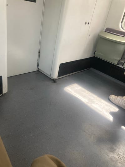 The image shows the interior of a public transportation vehicle, likely a train car, with a focus on the floor and lower walls. The floor is a speckled gray material, showing some wear and faint stains. A bright rectangular patch of sunlight illuminates the floor from the right side of the frame.

On the right side of the image, a person's lower legs and feet are visible, wearing white sneakers. The person is partially obscured by shadows and the bright sunlight.

The walls are white and appear to be made of a smooth, durable material. There are several features visible on the walls:
* On the left, a white door with a dark horizontal strip near the top is present. A thin vertical hinge is visible along its right edge.
* To the right of the door, a corner section of the vehicle's interior is visible. This section has a white, curved protrusion extending from the wall near the floor, featuring a black grate.
* Further to the right, a light green seat is visible, with purple upholstery. A black ribbed panel runs along the bottom of the wall behind the seat.

The overall impression is that of a functional, utilitarian interior space within a public transport system. The location is identified as Montcada i Reixac, Spain.