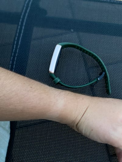A close-up, eye-level shot shows a person's left forearm and wrist wearing a green fitness tracker. The arm is light-skinned with visible fine hairs. The fitness tracker has a white face and a green band, which is loosely clasped around the wrist. The tracker is positioned over a dark, textured surface, possibly an outdoor chair or table, with a woven pattern. The background is out of focus, suggesting an outdoor setting. There are no other people or identifying features of the location visible in the image.