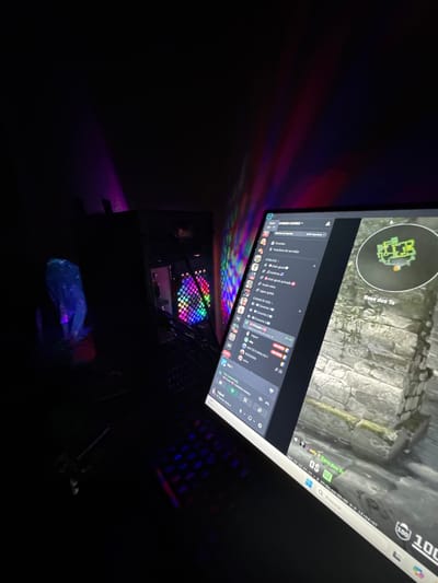 The image shows a dimly lit room featuring a computer setup. The monitor displays a screenshot of a Counter-Strike game, along with the Discord application. The screen is tilted upwards. Below the monitor, a computer case with RGB lighting is visible. A keyboard is partially visible to the right of the monitor. A plastic bag sits on the floor next to the computer. There is no evidence to indicate that the image was taken in Mirandela, Portugal.