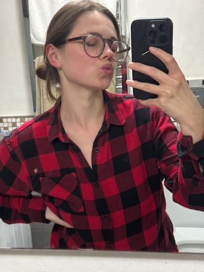 The scene depicts a mirror selfie taken by a female subject in a bathroom located in Cava de' Tirreni, Italy. The subject is shown from the mid-torso up, wearing a red and black buffalo plaid long-sleeved flannel shirt with the top button undone. Her hair is dark brown and pulled back in a bun. She wears round, dark-rimmed eyeglasses. Her eyes are closed, and her lips are pursed in a "kissing" gesture. Her right hand, holding a dark-cased smartphone (consistent with an iPhone featuring a three-lens camera system), is visible in the upper right portion of the frame, angled towards the mirror to capture the reflection. The subject's left hand is placed on her hip. The background behind the subject's reflection reveals a wall with a metal towel rack holding a white towel on the left, and a tiled wall featuring small, light-colored square tiles on the right. White bathroom fixtures are partially visible at the bottom edge of the frame.