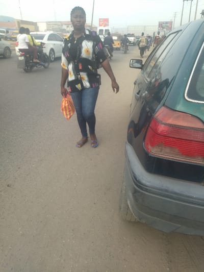 The image shows a woman in a black and white patterned shirt and blue jeans walking on a paved road in Way, Nigeria (latitude: 9.029, longitude: 7.584). She is carrying a yellow and red striped bag in her left hand. She is walking past a dark green car, and a man in a blue shirt is riding a motorcycle in the background. There are other cars and pedestrians in the distance, and buildings on either side of the road. The sky is overcast and the road is dusty.