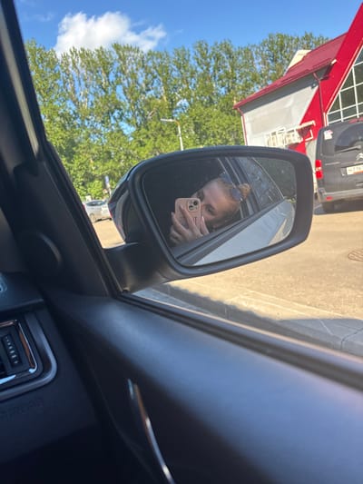 A woman is taking a selfie in her car's side mirror. She is looking at her phone, which is held in her right hand, and appears to be leaning out of the car window. She has her hair tied up and is wearing sunglasses on top of her head. The car interior, including part of the dashboard and door panel, is visible in the foreground. Outside the car, through the window and reflected in the mirror, there is a street scene with trees, buildings, and other vehicles. A black van with a license plate "M716" is parked on the right side of the road. A modern building with a red roof and grey facade is visible. The sky is blue with some white clouds. The image was taken in the city of Fedorino, Russia.