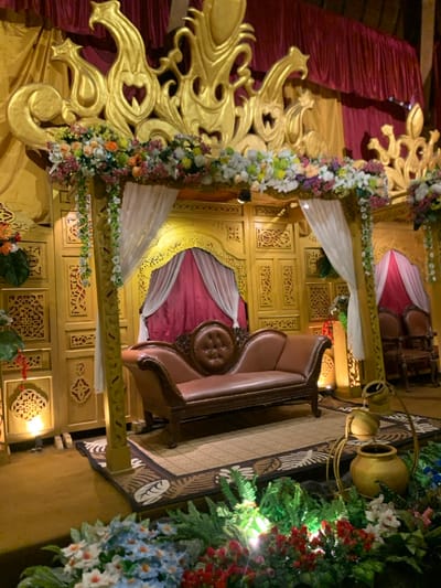 The image shows an indoor setting, likely a wedding ceremony or reception space, located at the coordinates -7.884 latitude and 112.525 longitude in the city of Batu, Indonesia. The scene features a wooden archway adorned with floral arrangements and white fabric draped over it. A  brown leather couch with a carved wooden frame sits on a mat in front of the archway. The wall behind the couch is made of intricately carved wood painted gold. A single  red curtain hangs  behind the couch.  The floor is carpeted with gold-colored fabric and patterned rugs.  The setting is illuminated by warm, artificial light. There are no people present.  There are flower arrangements in the foreground and a golden pot with a  cascade of water feature in the lower right corner of the frame.  The overall mood is one of celebration and festivity.