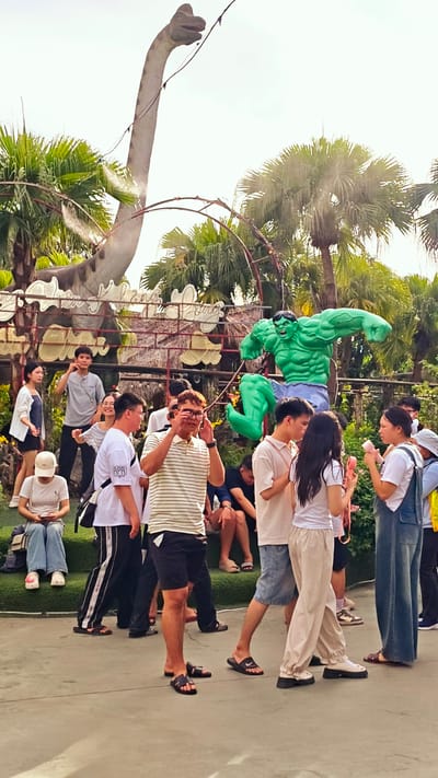 The image captures a lively scene at what appears to be an outdoor attraction, possibly a park or amusement area, in Hoa Lư Ward, Vietnam. In the background, a large, faded statue of a long-necked dinosaur with its mouth open stands tall against a bright sky, partially obscured by lush green palm trees. In front of the dinosaur, a vibrant, muscular green statue of the Hulk is frozen mid-leap, with his fists clenched.
A group of people, diverse in age and gender, are scattered throughout the foreground and middle ground, engaged in various activities. Some are posing for photos, others are looking at their phones, and a few are interacting with each other. A woman in a white t-shirt and blue jeans is sitting on a green tiered seating area, looking at her phone. A man in a striped shirt and shorts is standing and appears to be adjusting his glasses while looking towards the Hulk statue. To his right, a man and a woman are talking, with the woman holding a pink object. A woman in a denim jumpsuit is standing further to the right, also appearing to converse. The ground is a textured concrete surface, with some green artificial turf visible on the seating area. The overall atmosphere seems casual and summery, with people enjoying the attractions.