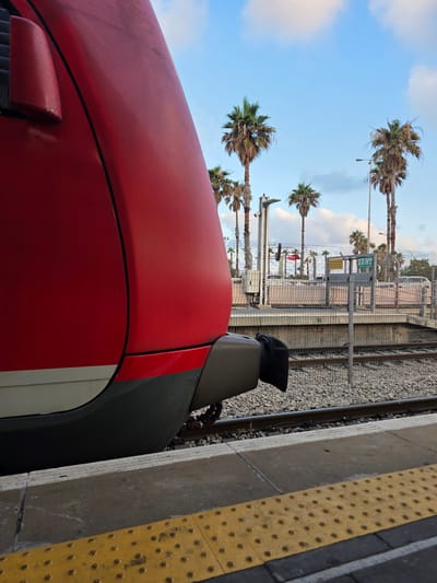 The image shows the front of a red train at a station platform in Haifa, Israel. The train is partially in frame on the left side, with its smooth, curved red exterior dominating the foreground. A dark grey buffer is visible at the front of the train. Beyond the train, a paved platform with a yellow tactile warning strip runs along the edge. Train tracks and ballast are visible to the right of the platform. In the background, a chain-link fence encloses the station area. Beyond the fence, several palm trees are prominent against a bright blue sky with scattered white clouds. A sign with Hebrew text is visible mounted on a pole within the fenced area, likely indicating station information. The overall scene depicts a typical urban train station environment.