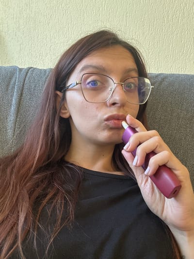 A young woman with long, dark brown hair and glasses is sitting on a couch, holding a pink electronic smoking device near her mouth. She is wearing a black t-shirt. The background consists of a textured, light yellow wall and a gray couch. There are no discernible features that place this image in Tryavna, Bulgaria.