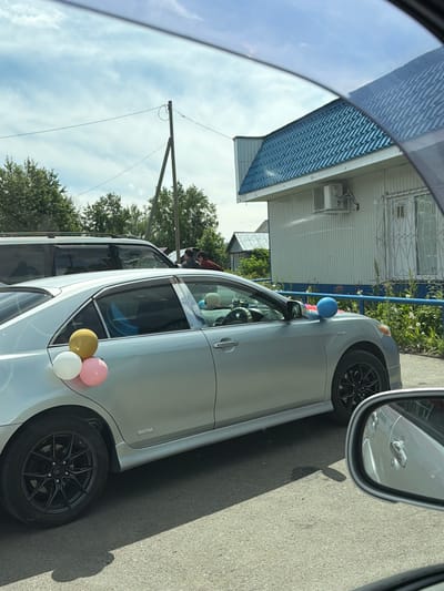 In Milkovo, Russia, a silver sedan is parked in front of a light-colored building with a blue metal roof. The car is decorated with three balloons on its rear passenger side: one white, one pink, and one yellow. Inside the car, a light blue balloon is visible on the passenger seat. The car has black rims on its wheels. In the background, trees and a utility pole are present. The sky is partly cloudy.