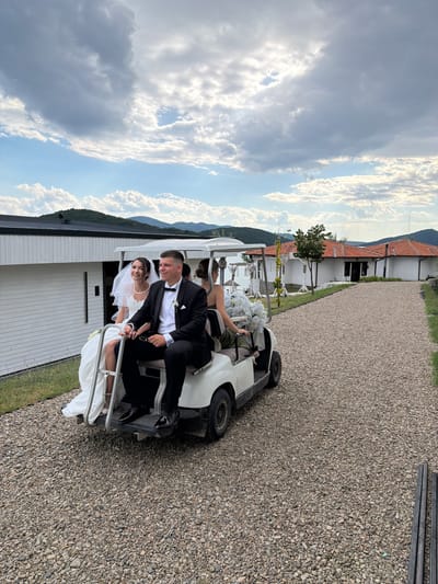 In Banya, Bulgaria, a wedding party is captured on a golf cart. The bride, in a white gown and veil, is seated next to the groom, who is in a black tuxedo with a bowtie. Two other women are also on the golf cart, one of whom appears to be a bridesmaid. The golf cart is driving on a gravel path. In the background, there are modern white buildings with dark roofs and traditional houses with red-tiled roofs. Mountains and a cloudy sky complete the scenic backdrop.