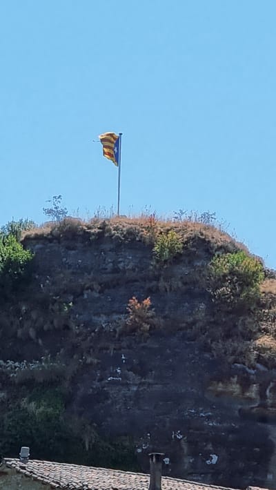 A flag, likely the Catalan flag due to its yellow and red stripes and blue square with a star, is prominently displayed on a flagpole atop a rugged, rocky hill. The flag appears to be flapping in the wind. The hill is covered in sparse vegetation, with some dry grasses and a few scattered trees. The sky above is a clear, bright blue. In the foreground, the roof of a building with red tiles is visible, along with parts of what appear to be chimneys or ventilation shafts. The overall scene suggests an outdoor setting, possibly in the rural area surrounding Rupit i Pruit, Spain, a region known for its dramatic natural landscapes and historic villages. The lighting indicates it is daytime, with the sun casting shadows on the terrain.