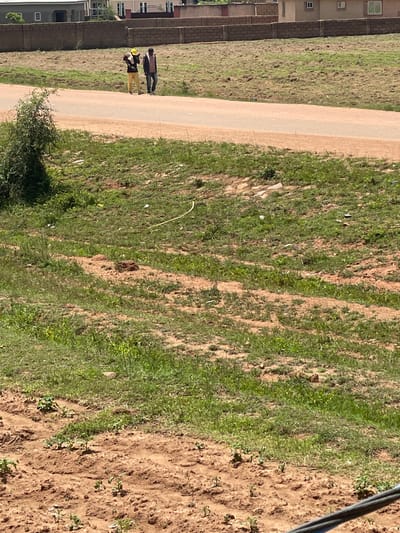 Two individuals are walking along a paved road, with one wearing a yellow hat and the other in a dark jacket. The scene is outdoors, likely in a semi-urban or rural area, as evidenced by the unpaved ground with vegetation and sparse buildings in the background. The foreground shows tilled soil with small plants emerging. The location is identified as Kwang, Nigeria.