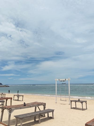 The image depicts a coastal scene in Monikeun, Indonesia. The sky is partly cloudy with shades of blue. In the middle ground, the turquoise ocean meets a sandy beach. Several wooden tables and benches are scattered across the sand. A white wooden swing set with the word "IMSURF" printed on the top is placed near the water's edge. Two individuals are standing near the shoreline. A large ship is visible on the horizon.