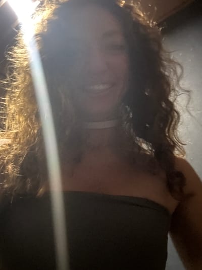 A person, likely female, is depicted in a portrait shot, presumably taken in Mù Cang Chải, Vietnam. A strong, bright light source from the upper left creates significant backlighting and lens flare, causing the left side of the subject's face and hair to be overexposed and appear golden, while the rest of their face and the background are cast in deep shadow. The subject has long, dark, curly hair and is smiling, revealing visible teeth. They are wearing a dark, strapless top and a white choker necklace. The background is uniformly dark and indistinct, lacking specific identifiable features.