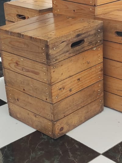 The image depicts multiple weathered wooden crates stacked on a tiled floor in Gadog, Indonesia. In the foreground, a stack of five light brown wooden crates is prominent, featuring visible nail heads with some white residue and rectangular cutout handles on their sides. The topmost crate exhibits a somewhat glossy surface, reflecting ambient light. To the right, a partial view of another similar stack of crates is visible. In the upper left background, part of a single wooden crate is present. The floor beneath the crates is composed of alternating dark (appearing as dark gray or black with white marbling) and plain white square tiles, forming a checkerboard pattern. No people or active interactions are visible in the scene.