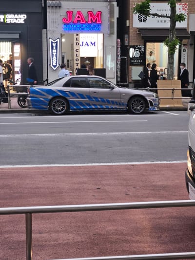The scene depicts a street in Shibuya, Japan. Notable establishments visible are "Grand Stage," "JAM Vintage & Select Used Clothing Store," and "Sakura Luggage Store Shibuya." People are present, some appearing to be pedestrians. A silver car with blue stripes is parked along the street. The environment includes a street with pedestrian crosswalks. The buildings appear to be multi-story structures.