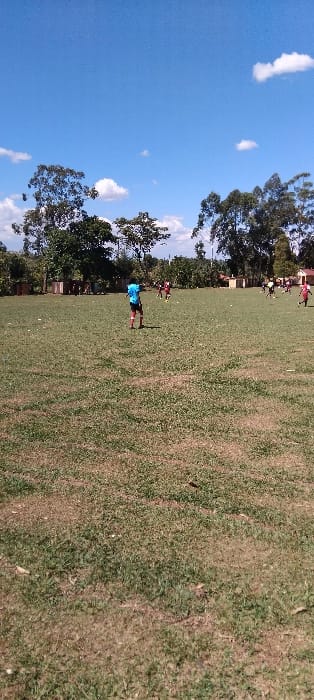 The image shows a grassy field with a group of people playing a game of soccer. There is a single person in blue wearing black shorts in the foreground and a few other people are further in the background. The sky is blue with some white clouds. This field appears to be in a residential area, evidenced by the presence of a few trees, a small building in the background, and a fence. The scene is set in Bungoma, Kenya at latitude: 0.554, longitude: 34.773.